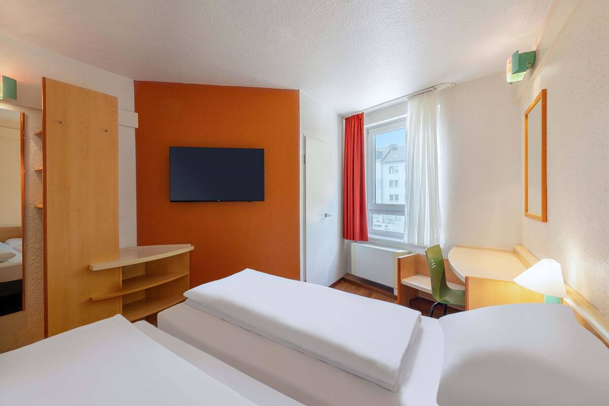 Ein Hotelzimmer mit Bett, Wandfernseher, Fenster mit Vorhängen und einem Sessel.