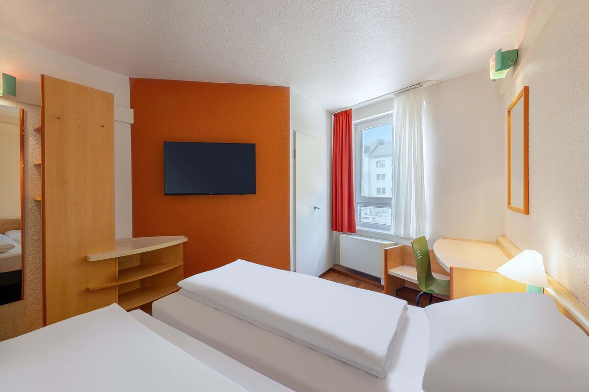 Une chambre d’hôtel avec un lit, un téléviseur fixé au mur, une fenêtre avec des rideaux et une chaise.