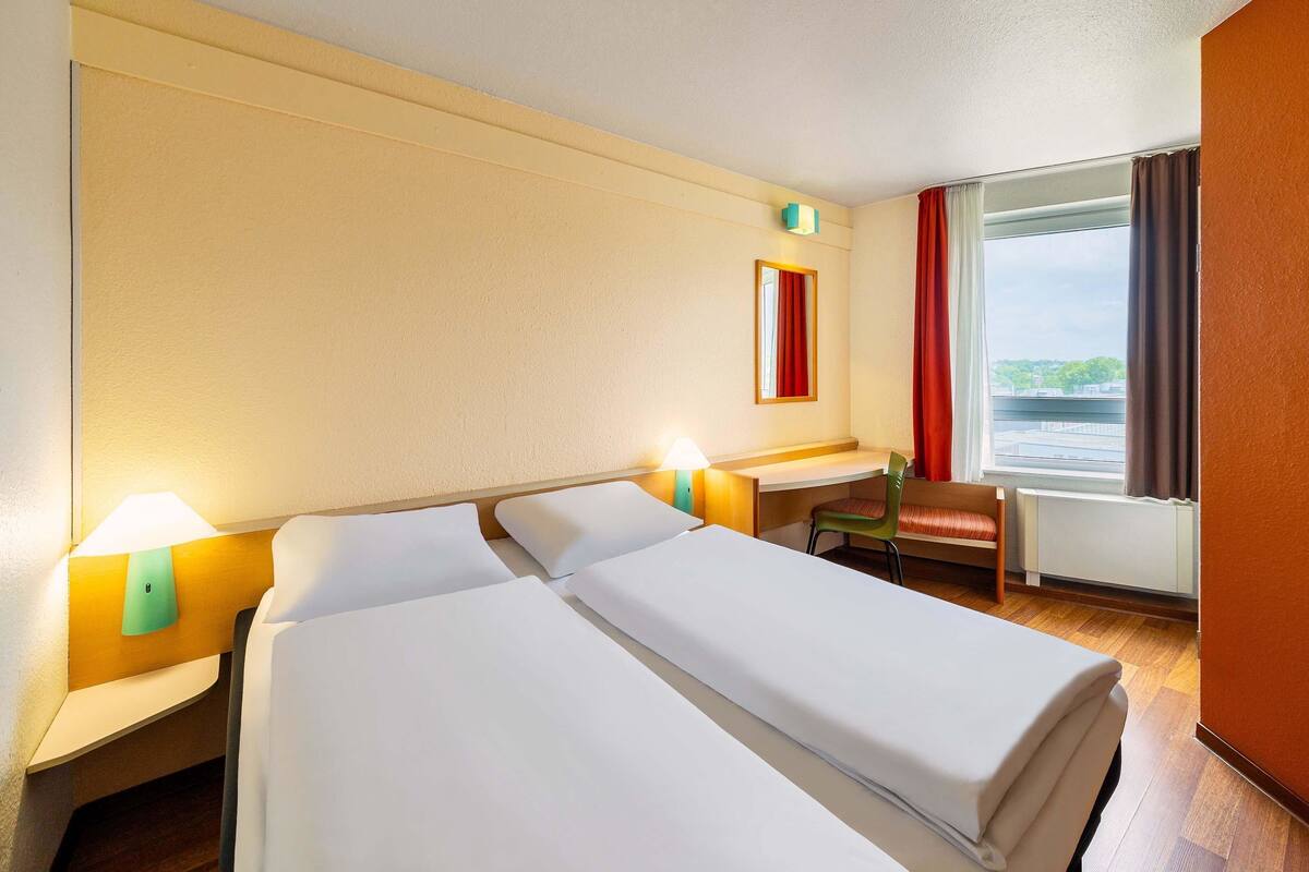 Ein Hotelzimmer mit zwei Betten, einem Schreibtisch, einem Stuhl, einem Fenster mit Vorhängen und einer Lampe.