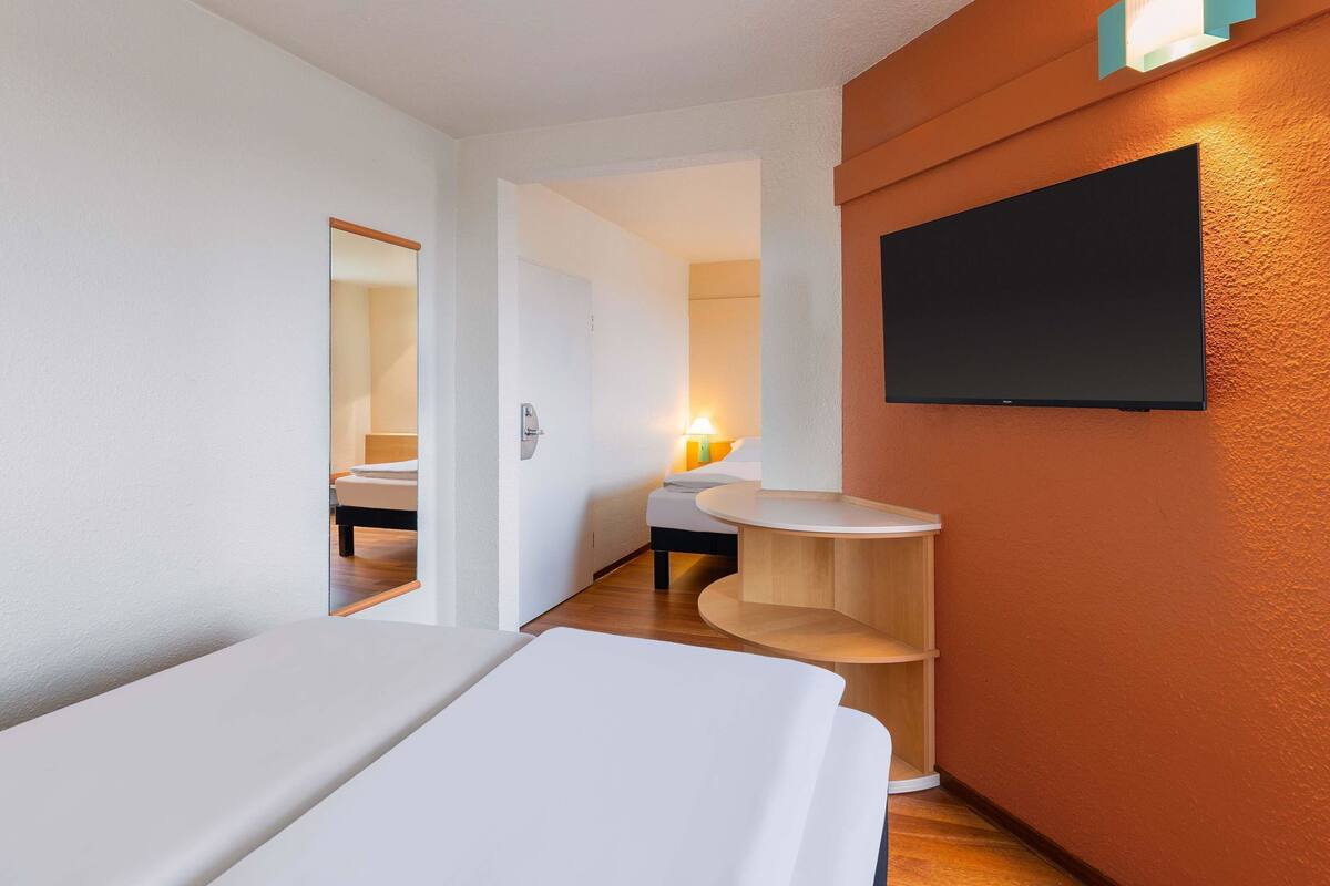 Ein Hotelzimmer mit einem Bett, einem kleinen Holztisch, einem an der Wand befestigten Fernseher und einer Tür zu einem anderen Raum.