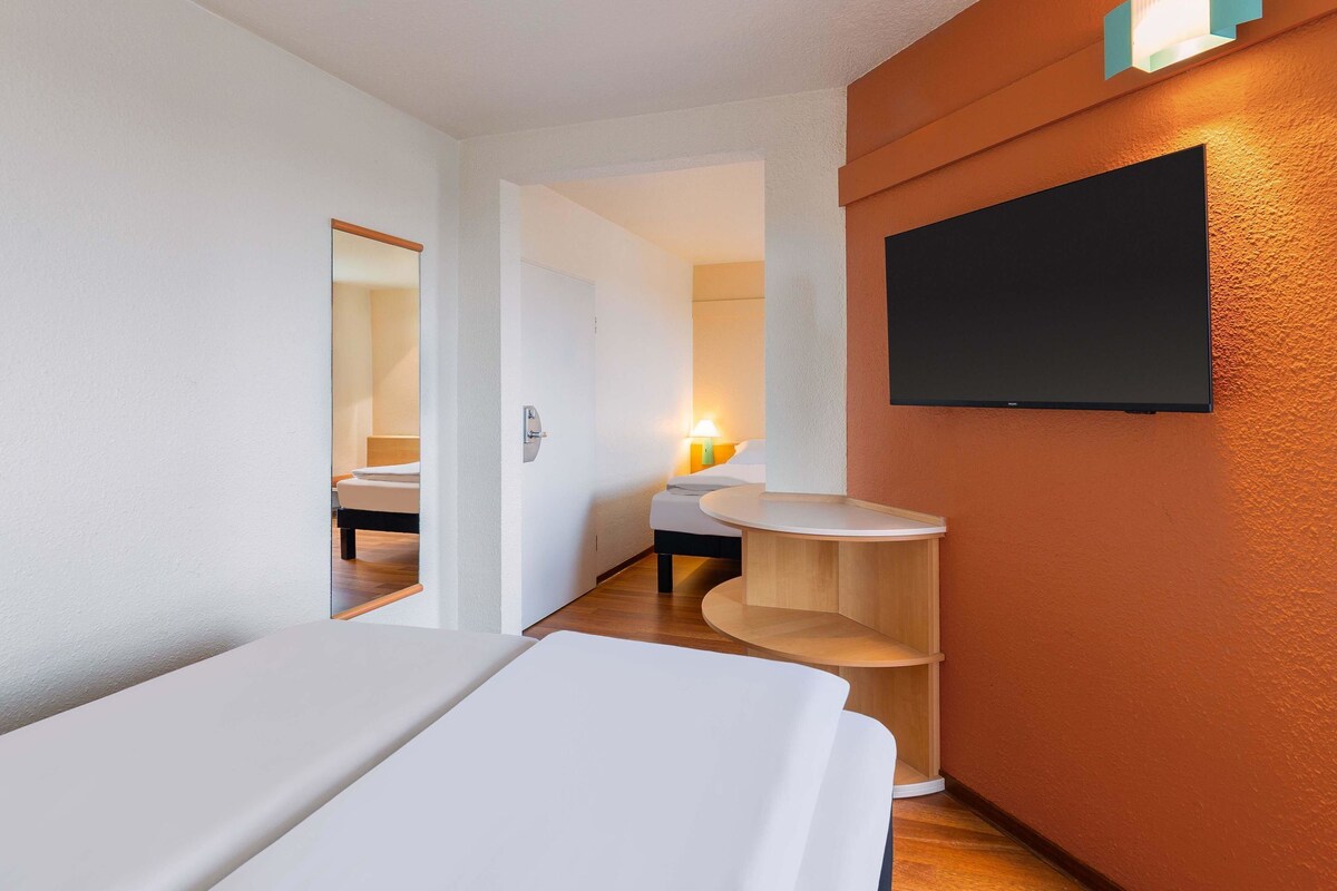 Une chambre d’hôtel avec un lit, une petite table en bois, un téléviseur fixé au mur et une porte donnant sur une autre pièce.