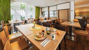 A modern dining area with wooden tables and chairs, large windows with green curtains, and a kitchen visible in the background. - B&B HOTEL Bochum Hbf-Süd (Bochum)