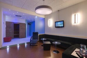 A modern lounge area with a sofa, a chair, a small table, and a television mounted on the wall. - Best Western Hotel Ambassador (Baunatal)