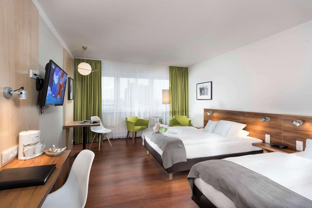 Habitación de hotel con dos camas, un escritorio con cafetera, televisor montado en la pared y vista a la ciudad a través de amplias ventanas.