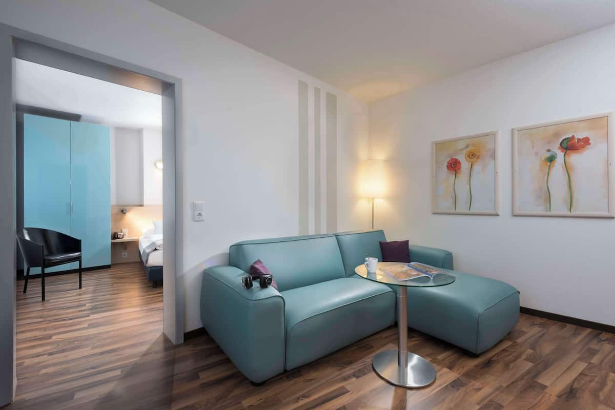 Una sala moderna con un sofá color turquesa, una mesita redonda y una lámpara. Hay dos cuadros en la pared, una cama al fondo y piso de madera.