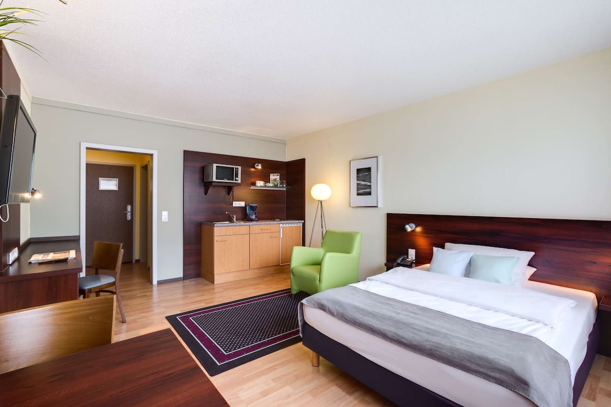 Habitación de hotel con cama, escritorio, silla, kitchenette y televisor de pantalla plana.