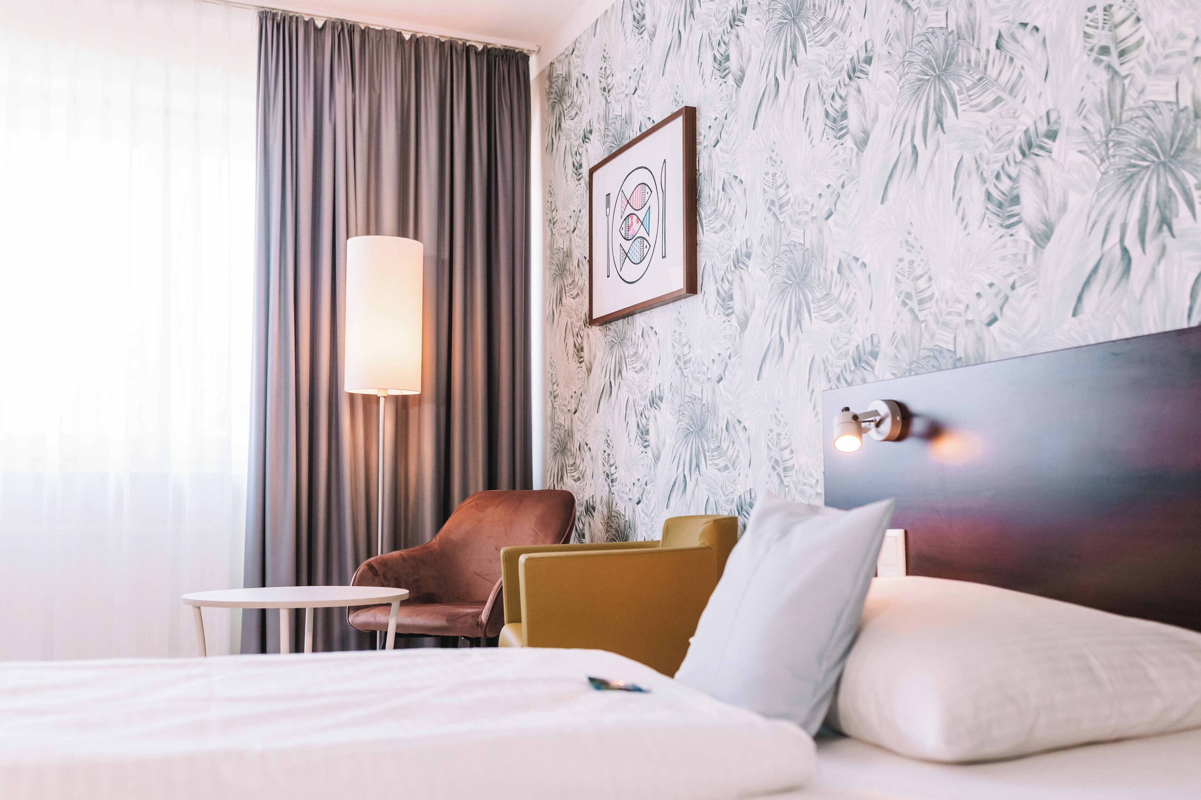 A hotel room with a bed, a chair, a table, a lamp, and a wall with a tropical wallpaper.