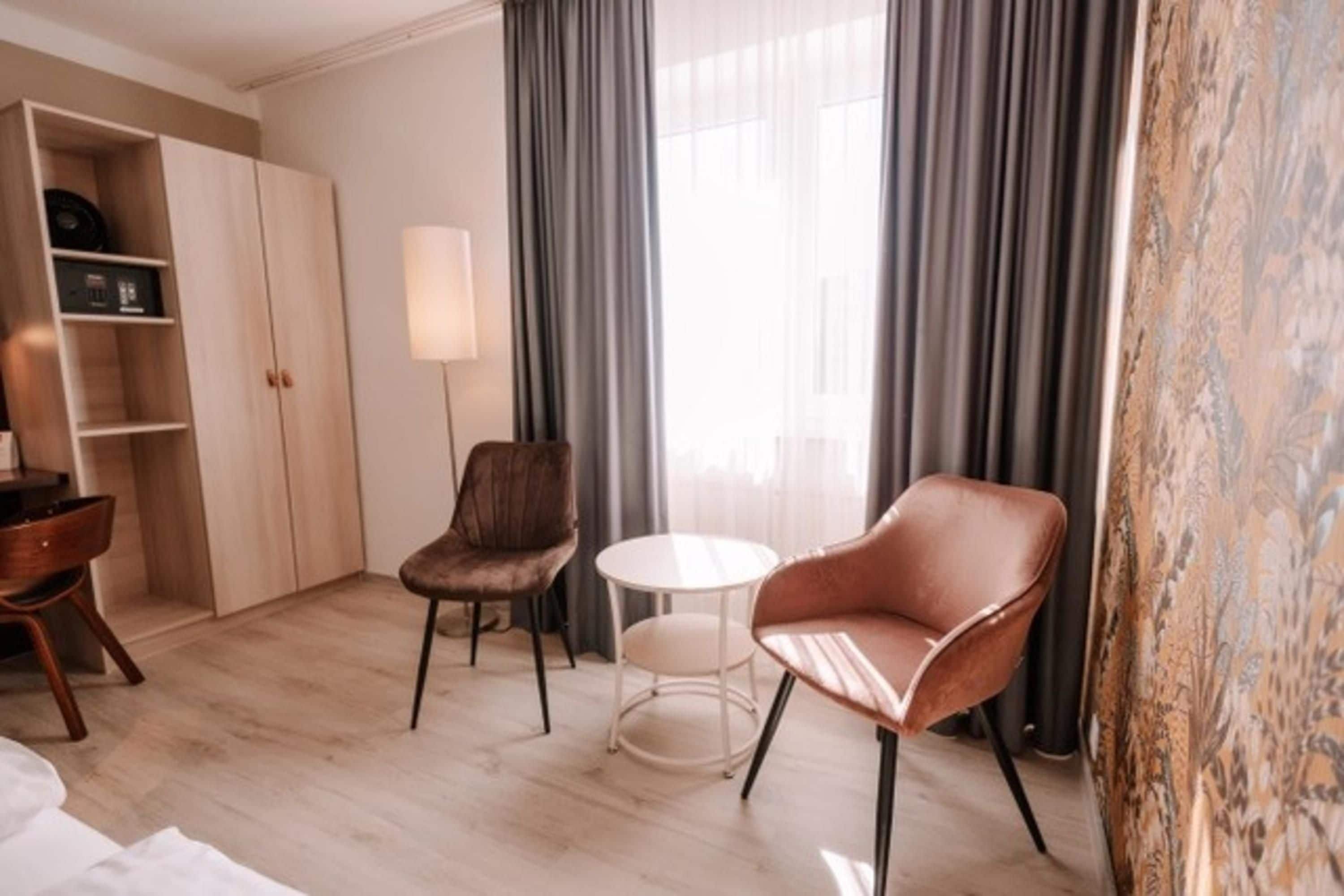 Ein Hotelzimmer mit einem Holzschrank, einem braunen Ledersessel, einem weißen runden Tisch und einem großen Fenster mit Vorhängen.