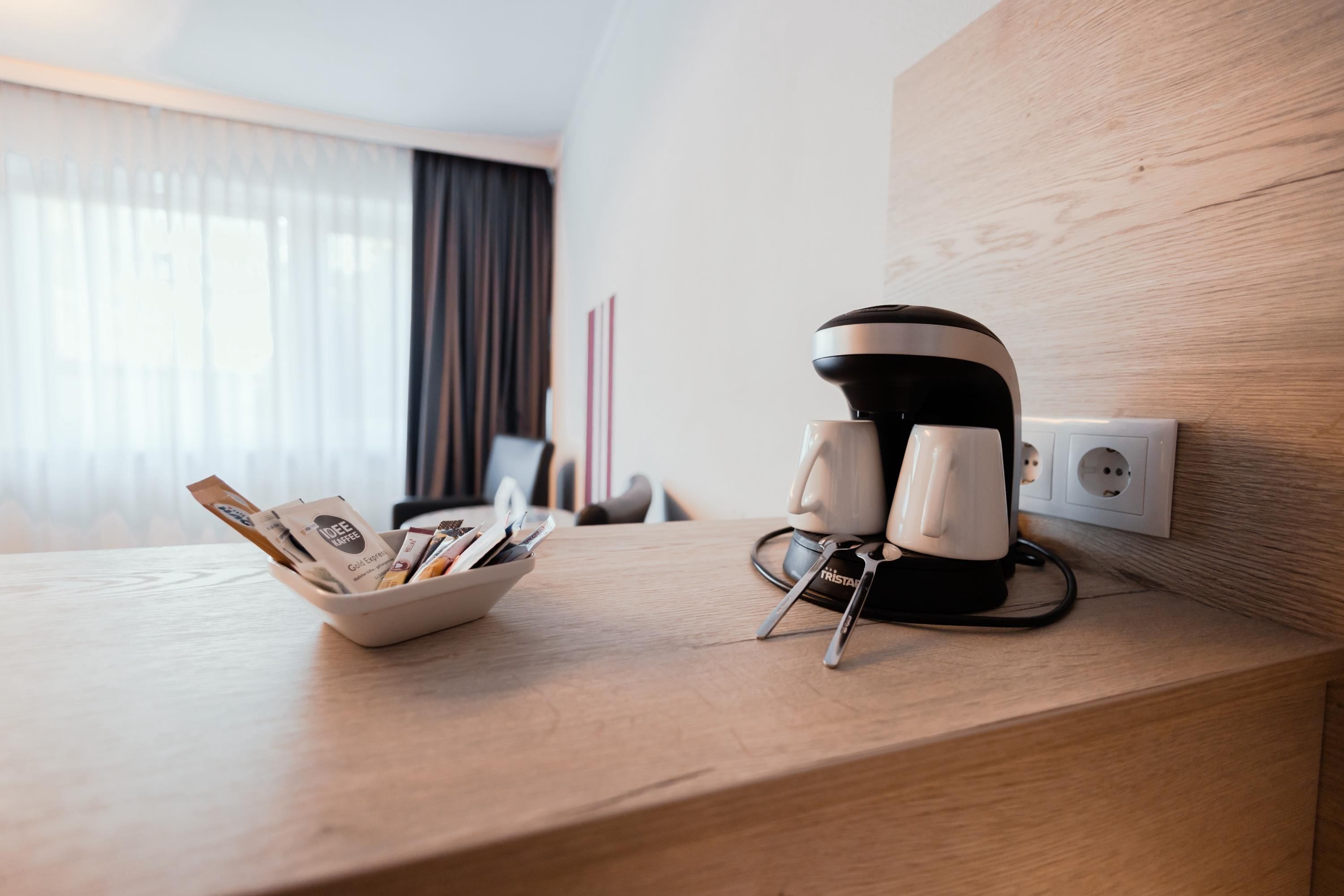 Ein Hotelzimmer mit einem Holztisch, einer Kaffeemaschine, zwei Tassen und einer Schale mit Kaffeepäckchen.