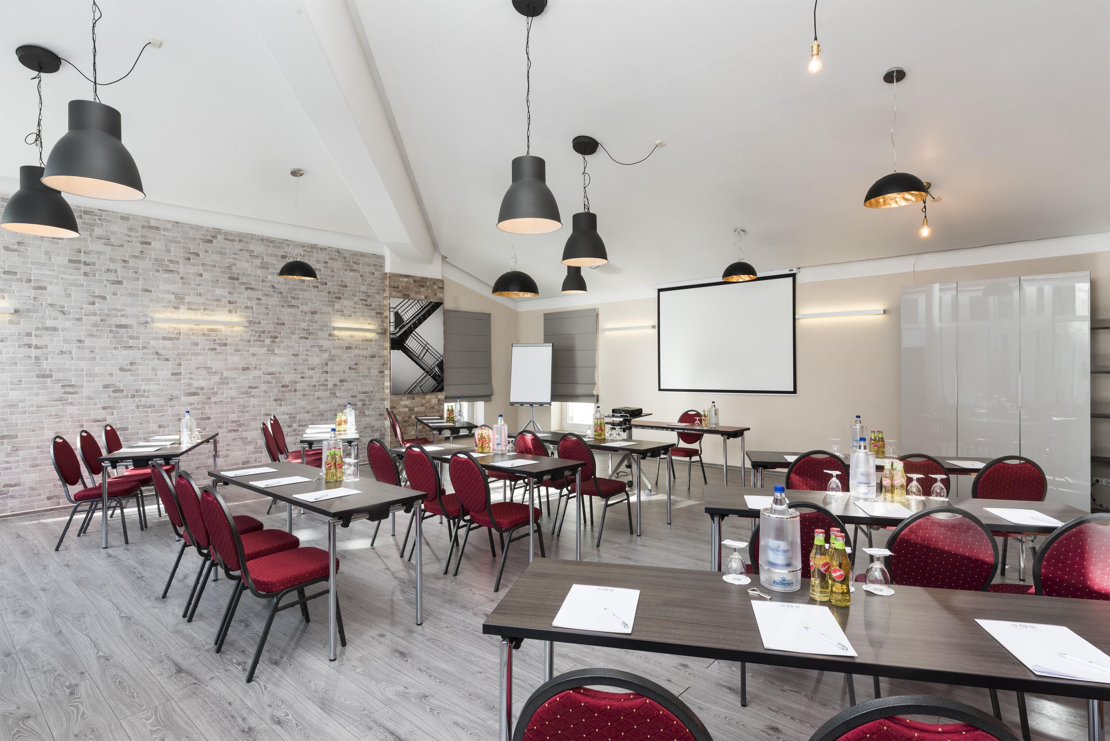 A conference room with multiple tables, red chairs, black pendant lights, a whiteboard, and a projector screen.