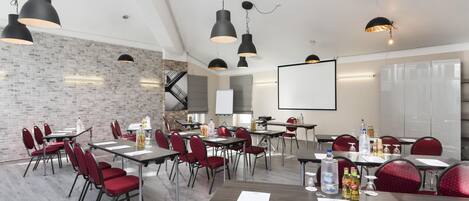 A conference room with multiple tables, red chairs, black pendant lights, a whiteboard, and a projector screen.