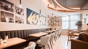 A modern restaurant interior with a long bench seating area, multiple tables, and a large abstract painting on the wall. - Best Western Hotel Ambassador (Baunatal)