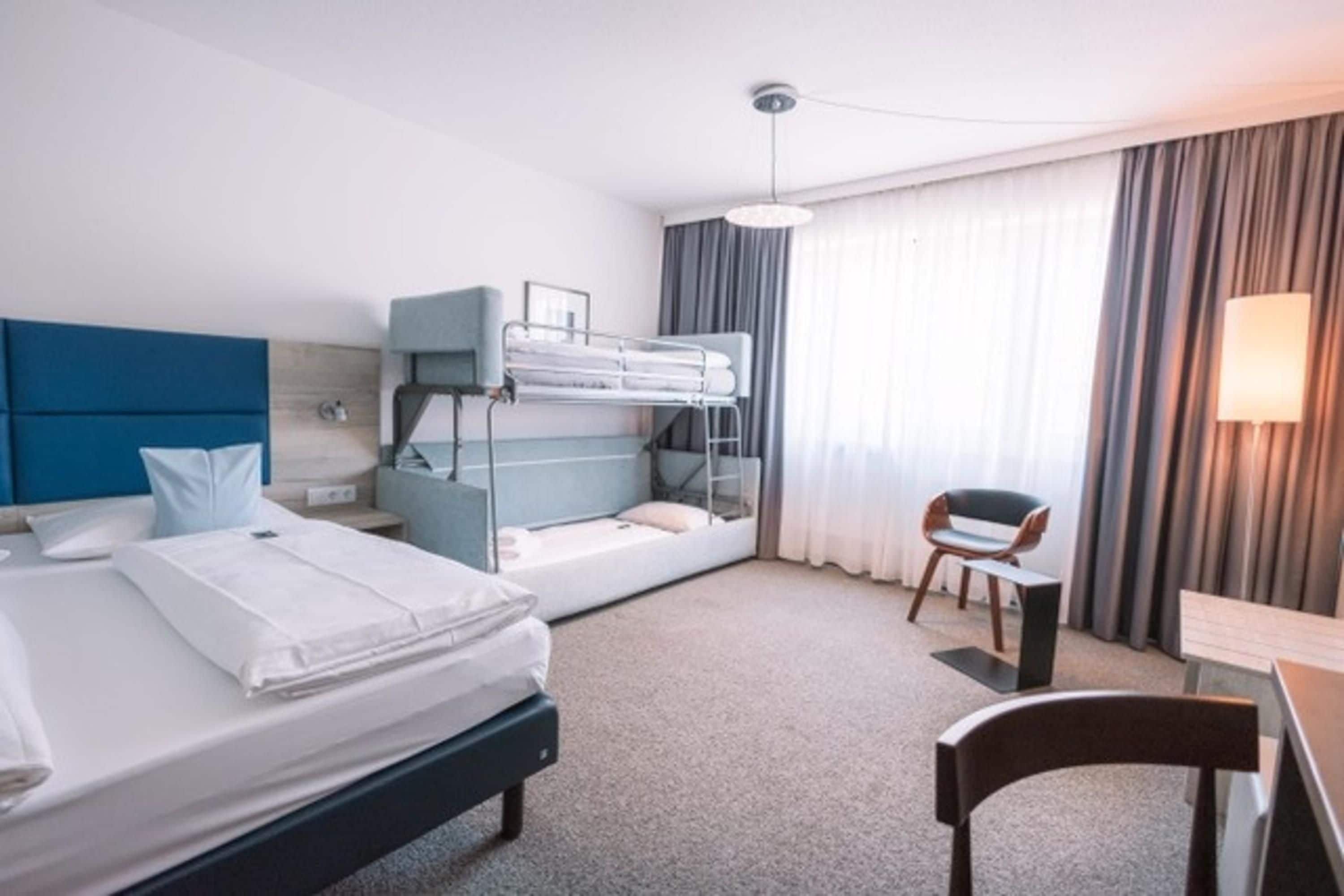 Habitación de hotel con literas, un escritorio, una silla, una lámpara y un ventanal grande con cortinas.