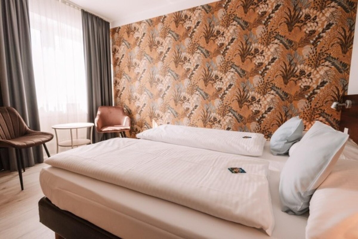 Habitación de hotel con una cama grande, una mesita redonda pequeña, una silla y un papel tapiz estampado.