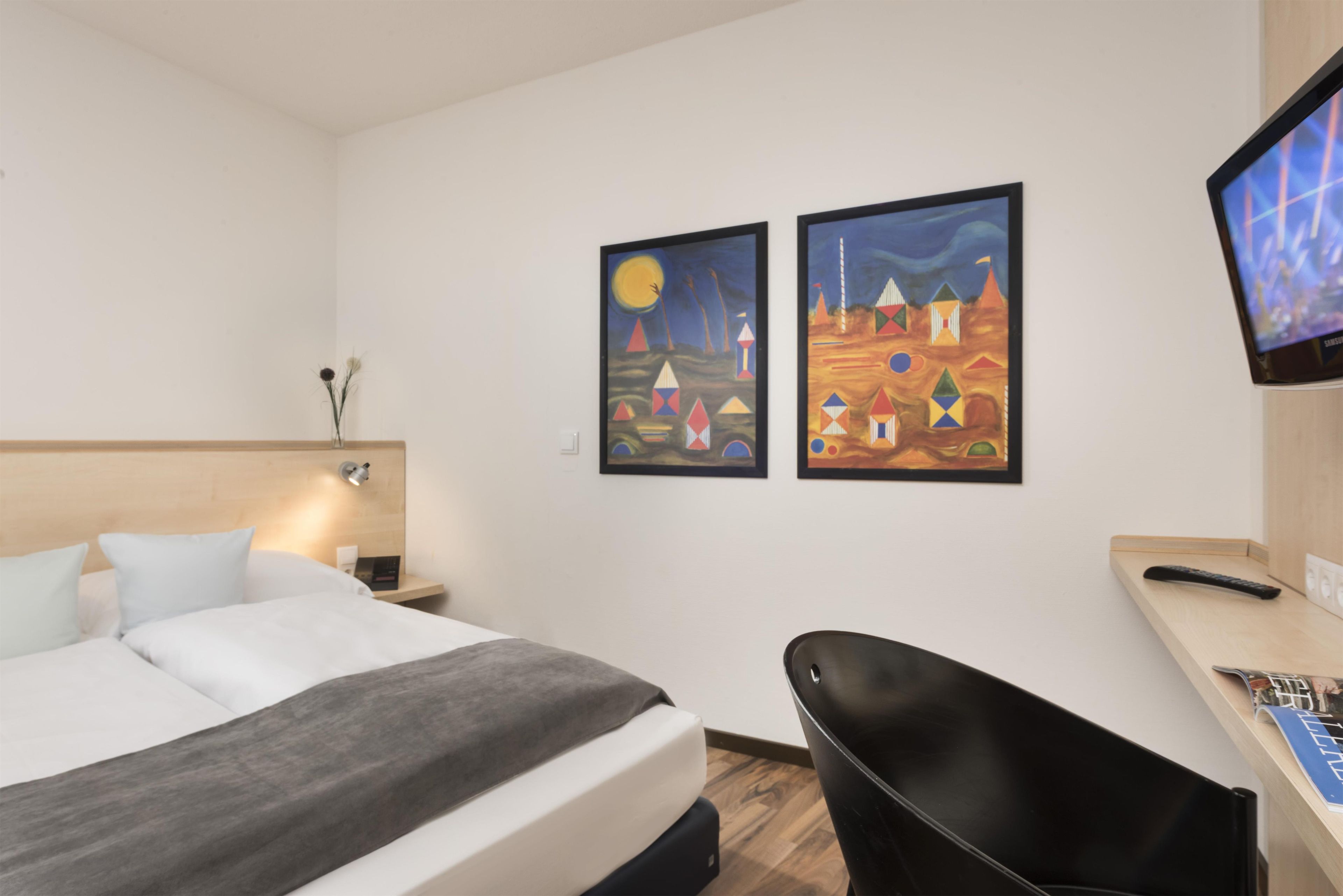 A hotel room with a bed, a desk with a computer, a chair, and two paintings on the wall.