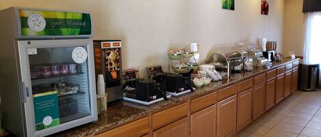 Zona de desayuno tipo bufé con una vitrina refrigerada, máquinas de café y varios recipientes de comida.