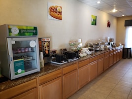 Zona de desayuno tipo bufé con una vitrina refrigerada, máquinas de café y varios recipientes de comida.