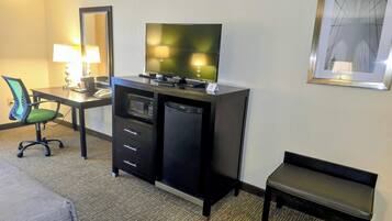 Habitación de hotel con escritorio, silla, cama, televisión, refrigerador minibar y un banco.