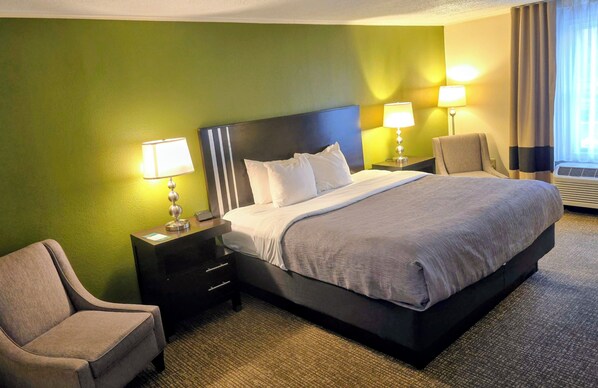 Quarto de hotel com uma cama grande, duas mesas de cabeceira com luminárias, uma cadeira e piso coberto por carpete.