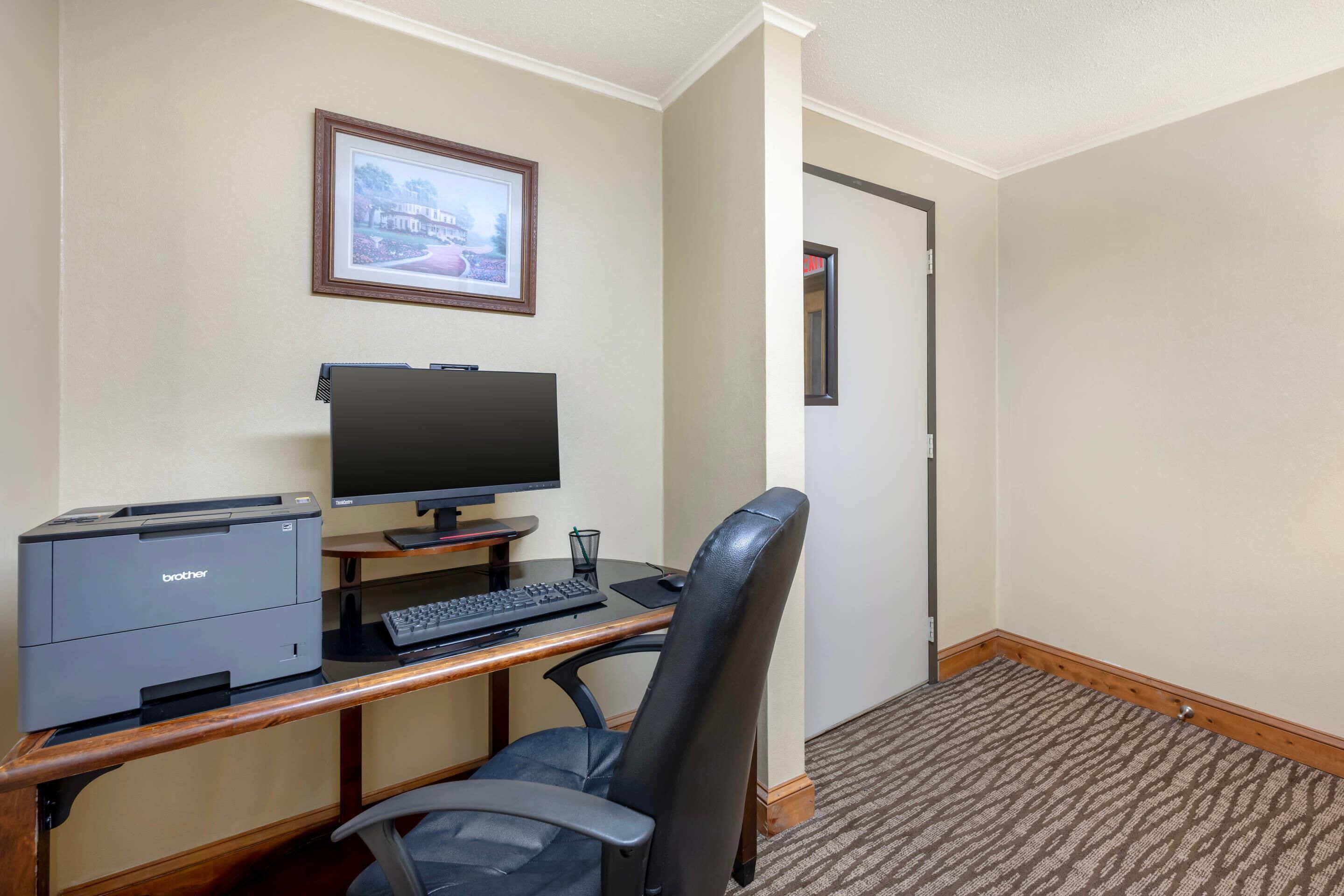 A hotel room with a desk, a computer, a printer, a chair, and a framed picture on the wall.