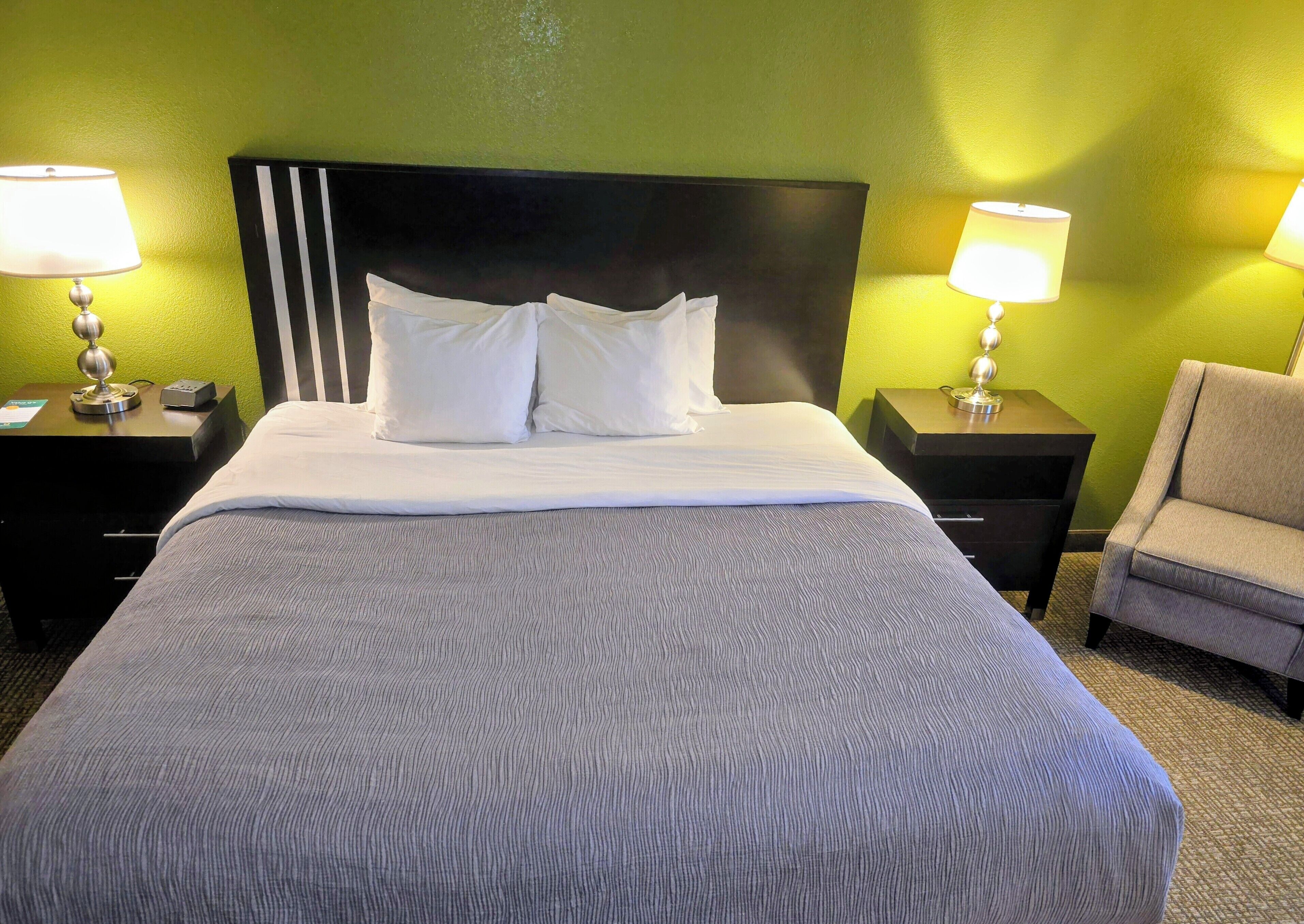 A neatly made bed with white linens, two bedside lamps, a black headboard, and a grey armchair.