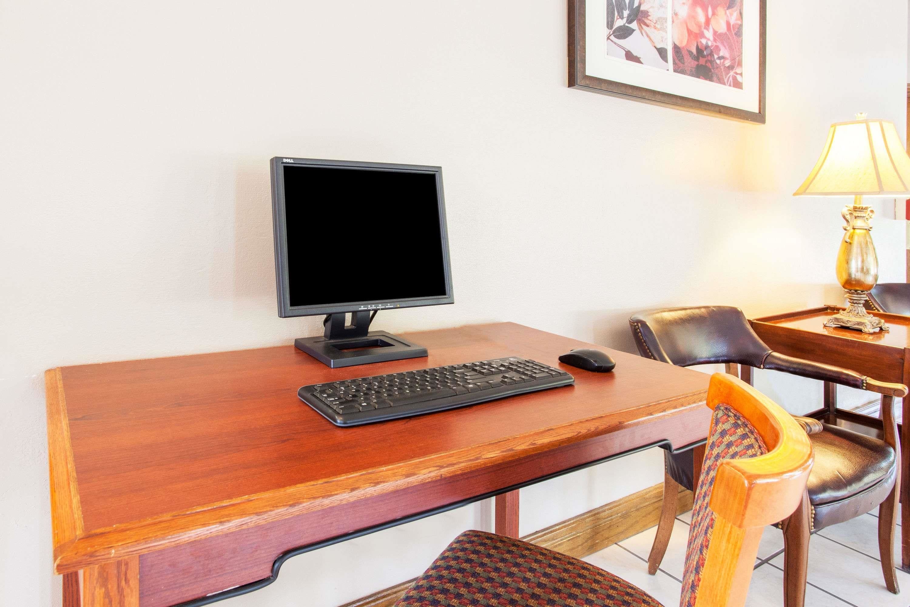 Ein Holztisch mit einem Computermonitor, Tastatur und Maus. Dazu ein Stuhl mit Sitzfläche aus Gewebe und eine Lampe auf einem Tisch daneben.