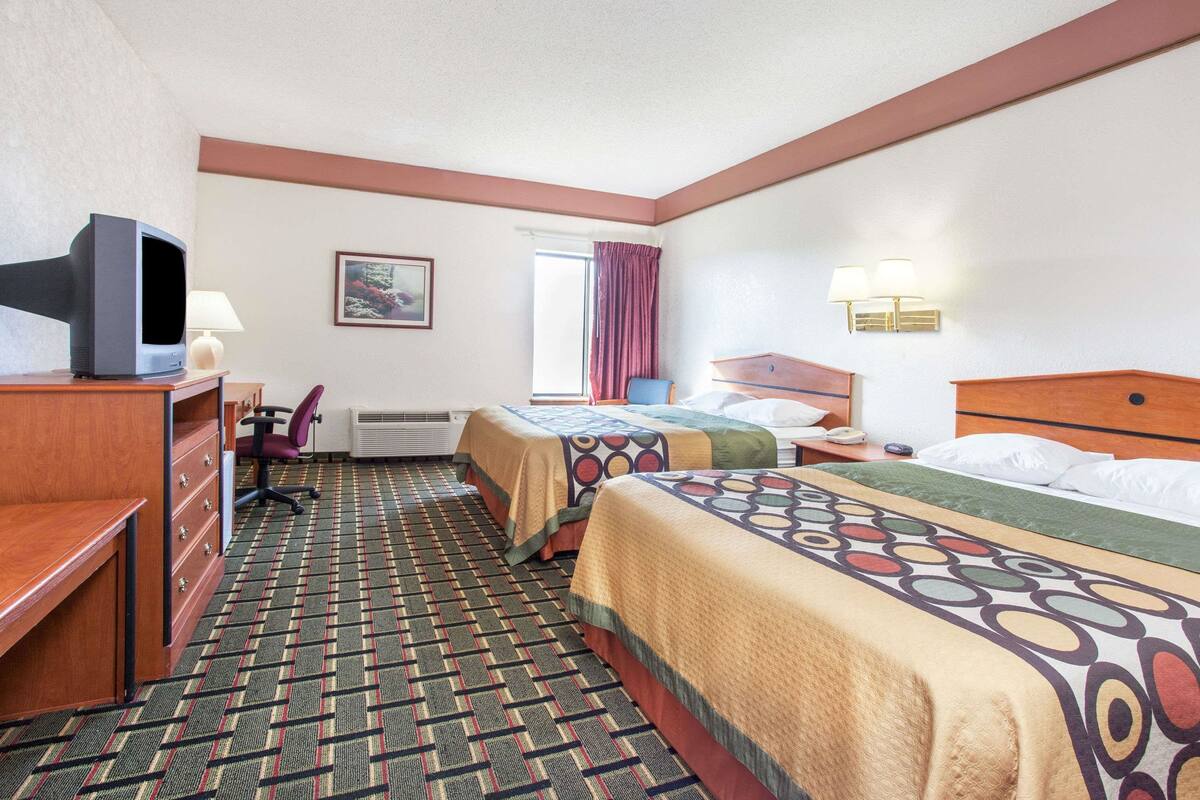 Habitación de hotel con dos camas, un escritorio con silla, televisor, ventana con cortinas y lámpara de pared.