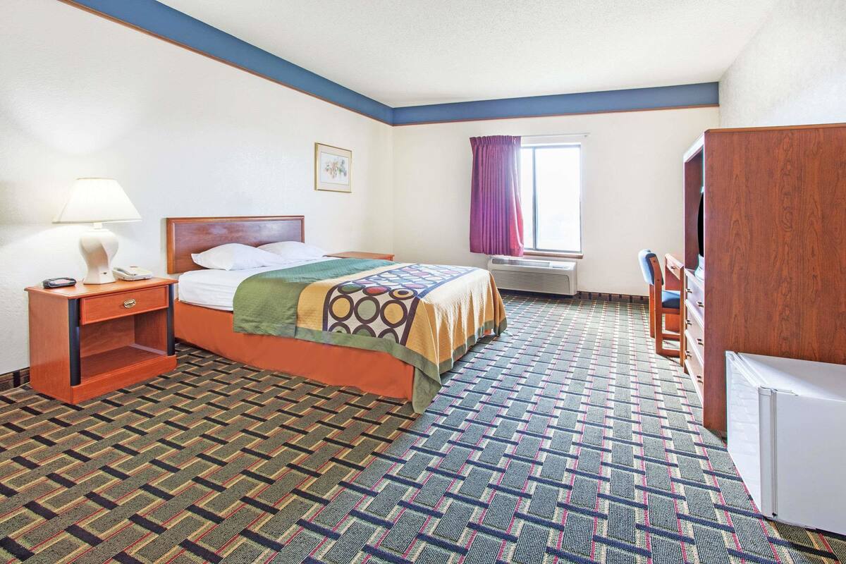 Habitación de hotel con cama, mesita de noche, escritorio, silla, ventana con cortinas y radiador.