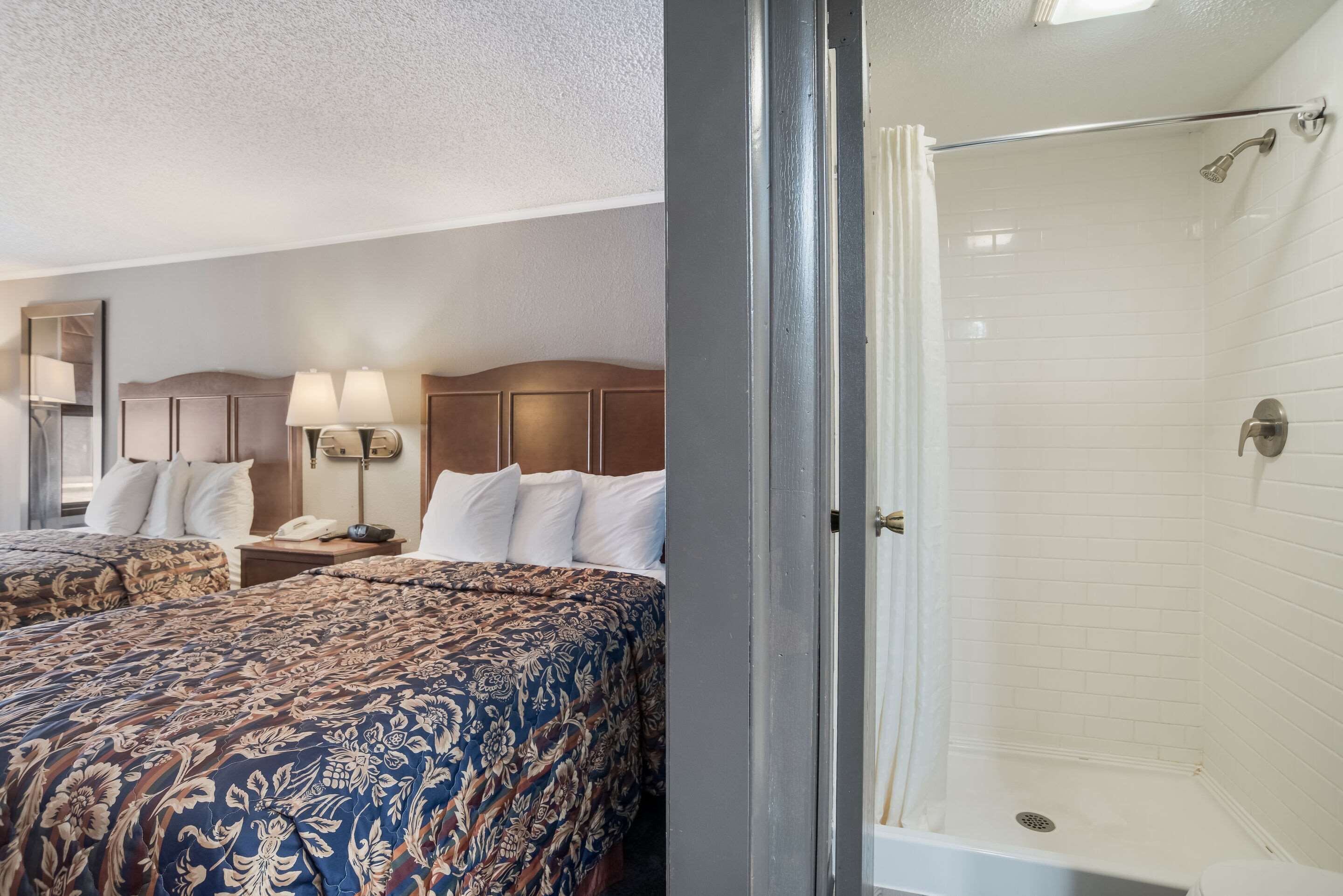 A hotel room with two beds, a bedside table with lamps, a telephone, and a shower area with a glass door.
