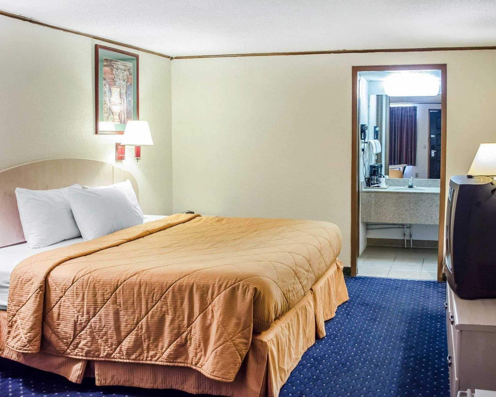 Habitación de hotel con cama, escritorio con televisor, baño visible a través de una puerta abierta y un cuadro en la pared.