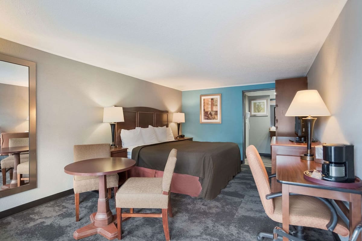 A hotel room with a bed, a desk with a lamp, a chair, a coffee maker, and a mirror.