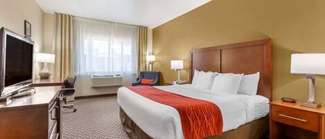 A hotel room with a large bed, a desk with a TV, a chair, a desk lamp, a bedside table with a lamp, and a window with curtains.