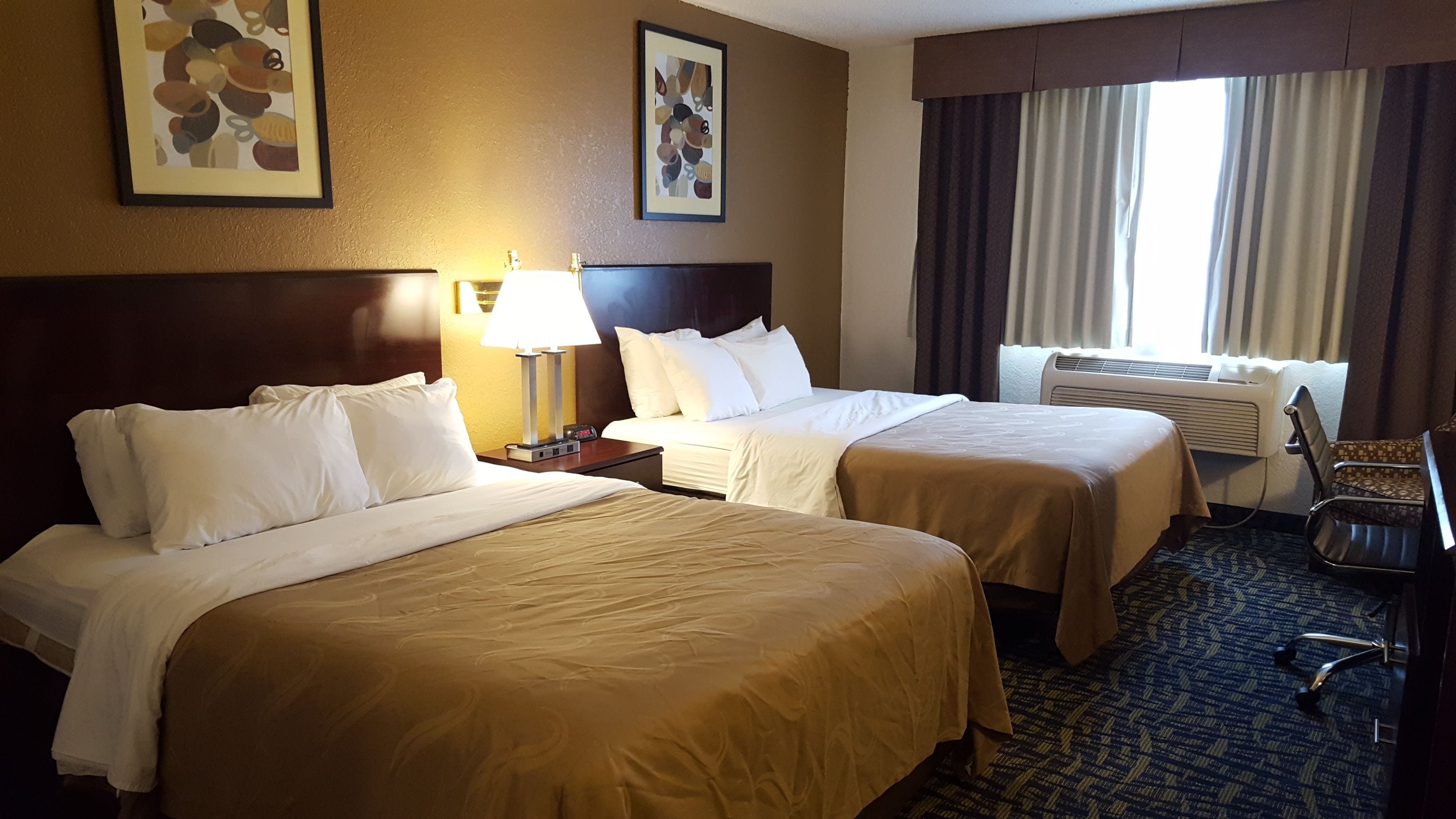 A hotel room with two beds, a desk with a lamp, a chair, and a window with curtains.
