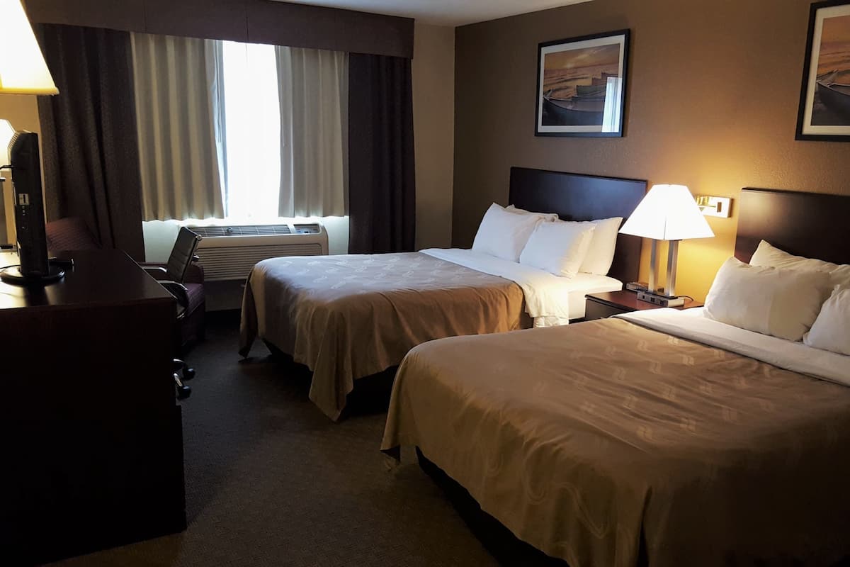 Een hotelkamer met twee bedden, een bureau, een stoel, een bureaulamp, een nachtkastje, een lamp, een raam met gordijnen, een airconditioning en een lijst aan de muur.