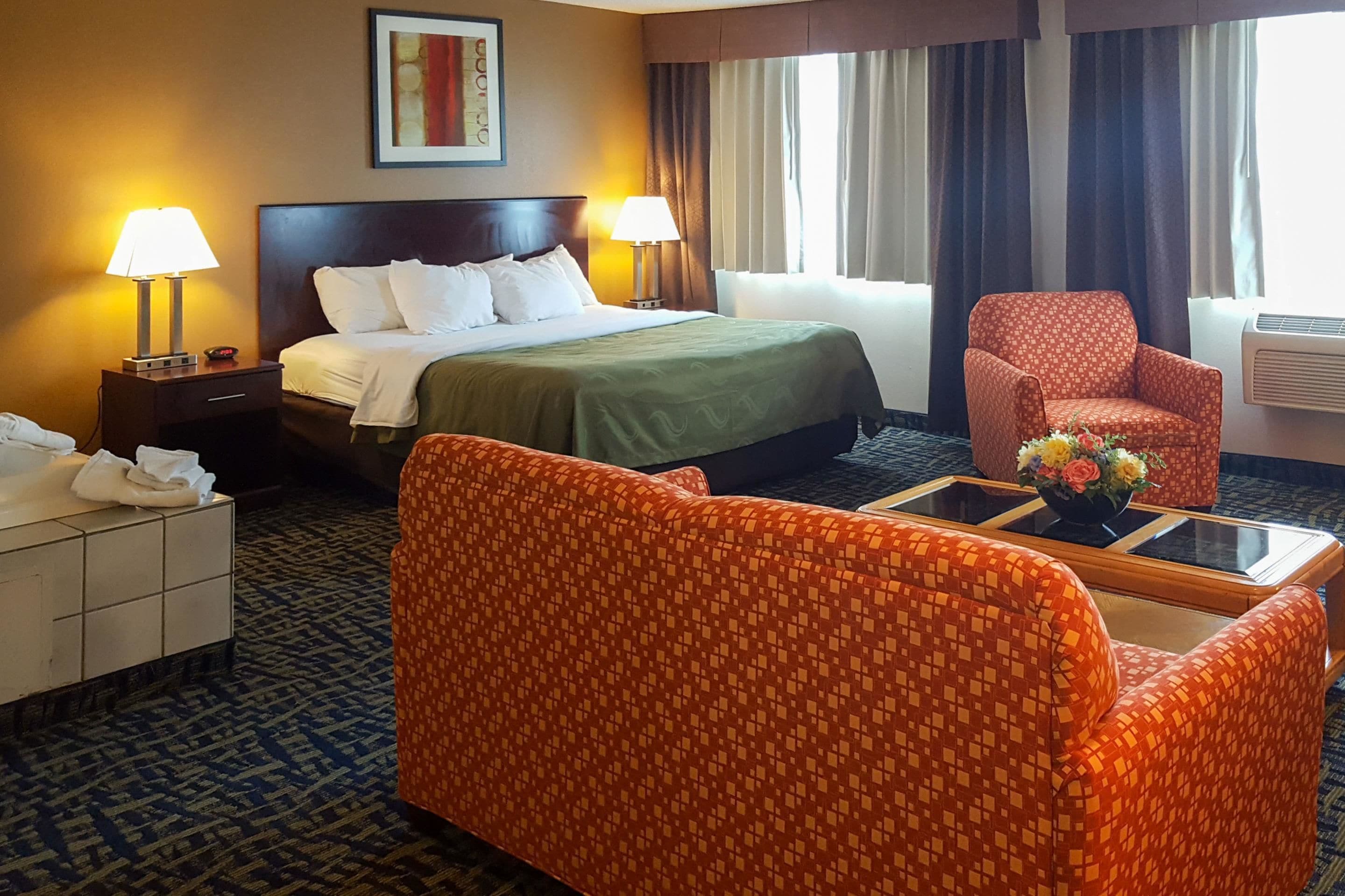 Ein Hotelzimmer mit einem Bett, zwei Nachttischlampen, einem Sessel, einem kleinen Tisch mit Blumenvase und einer Wand-Klimaanlage.