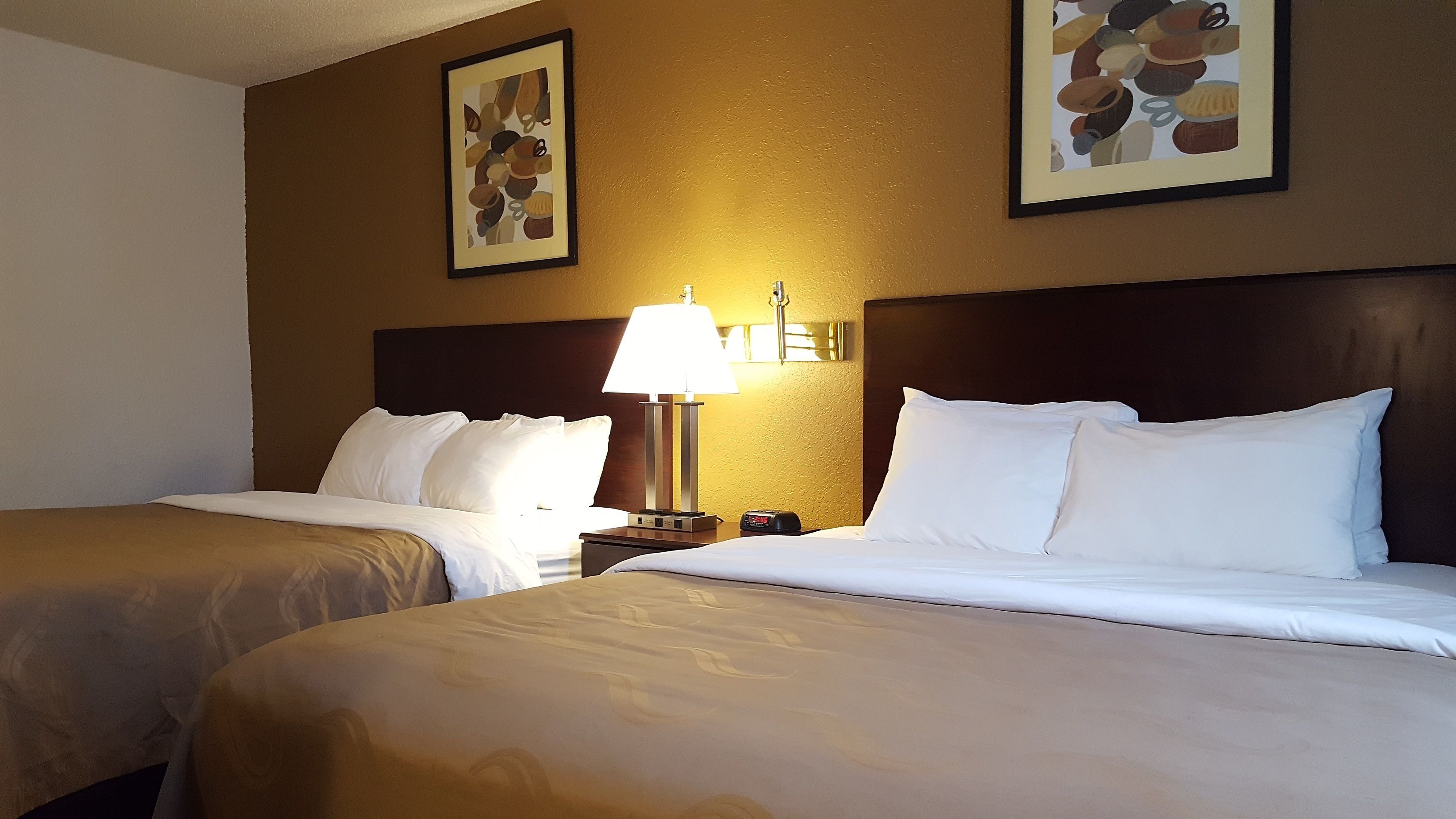 A hotel room with two beds, a nightstand with a lamp, and two framed pictures on the wall.