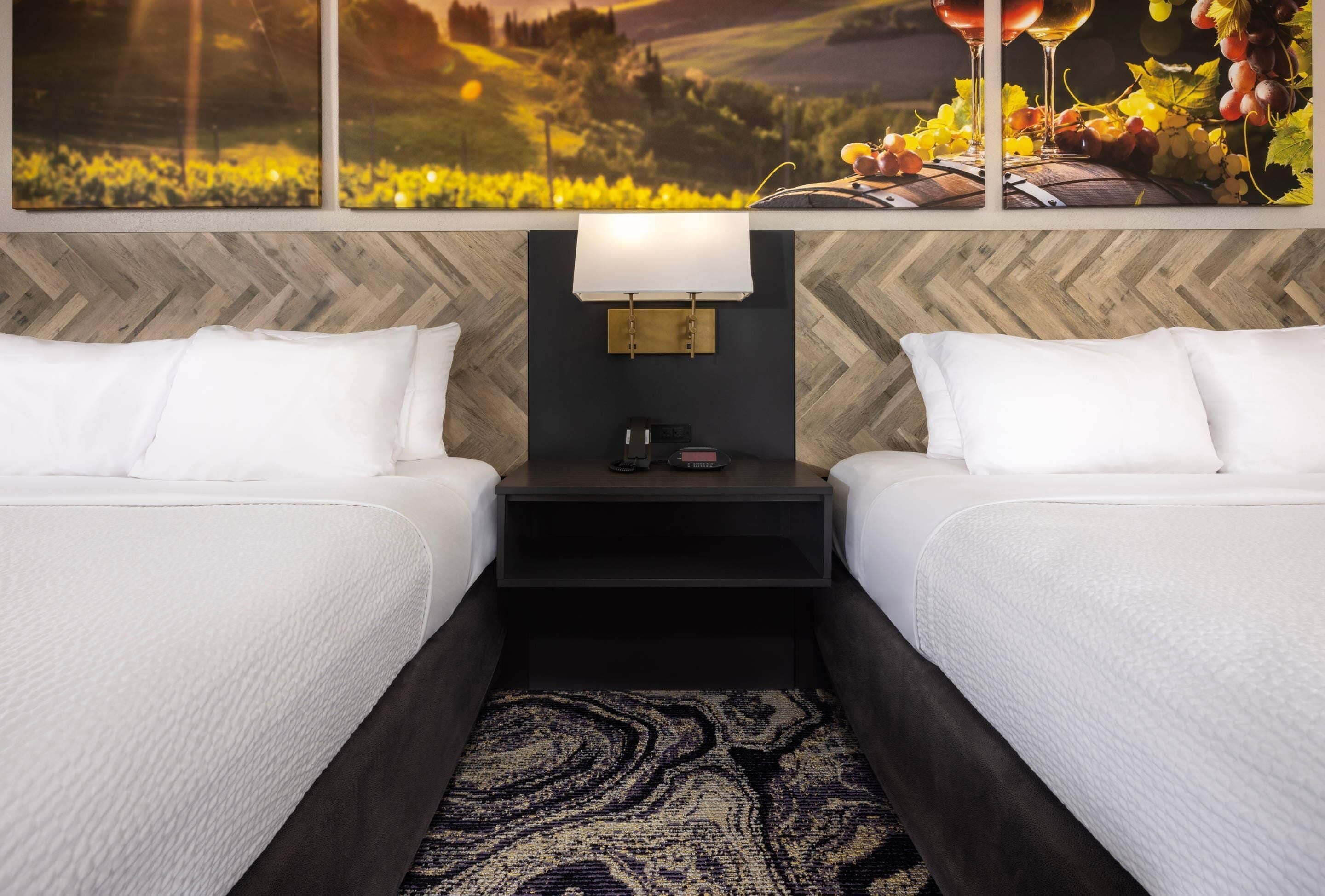 A hotel room with two beds, a nightstand with a lamp, and a large window with a vineyard view.