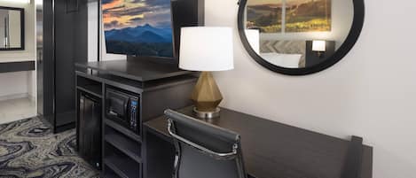 A hotel room with a flat-screen TV, a desk with a lamp, a chair, a coat rack, a microwave, and a large mirror.