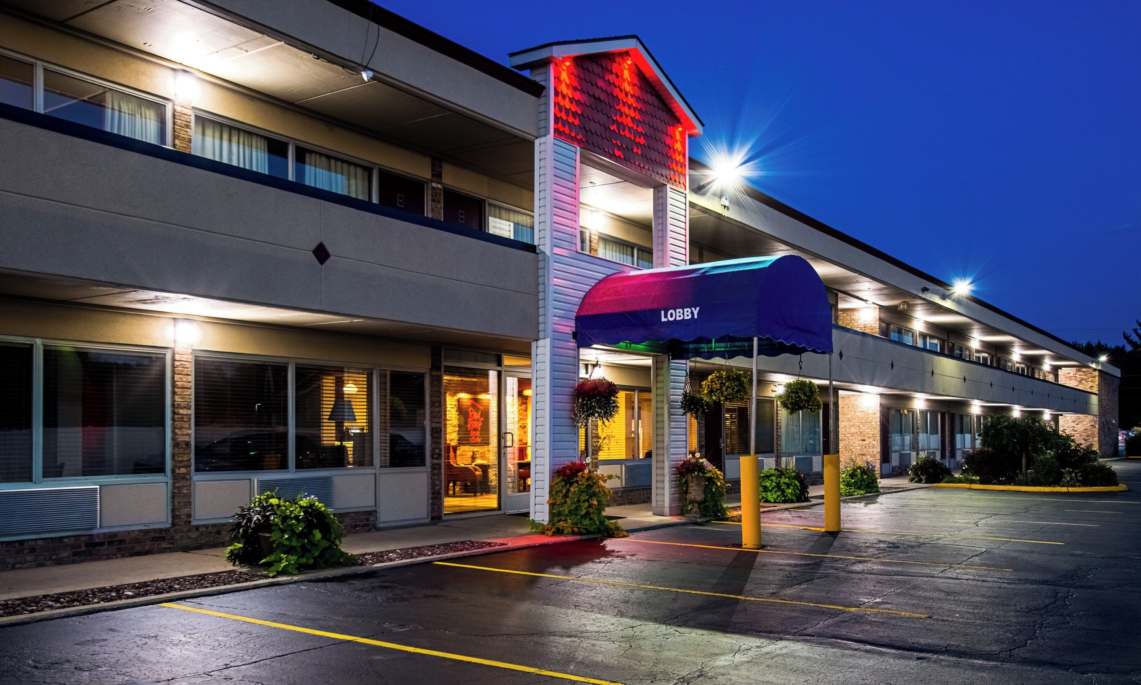 A hotel with a prominent red and white sign, a covered entrance with a blue awning, and a parking lot.