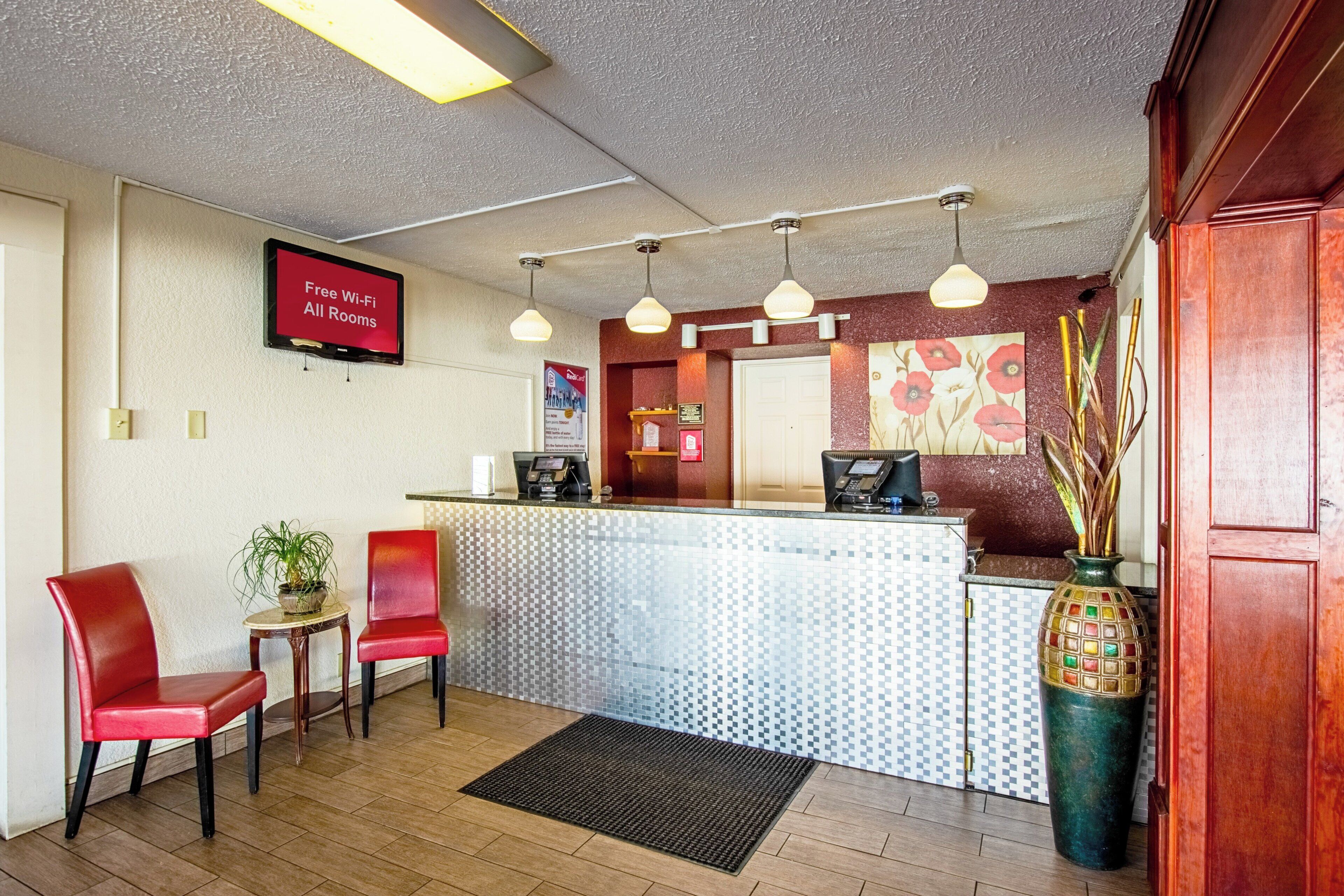 A reception area with a flat-screen TV, a reception desk, two red chairs, a potted plant, and a decorative vase.