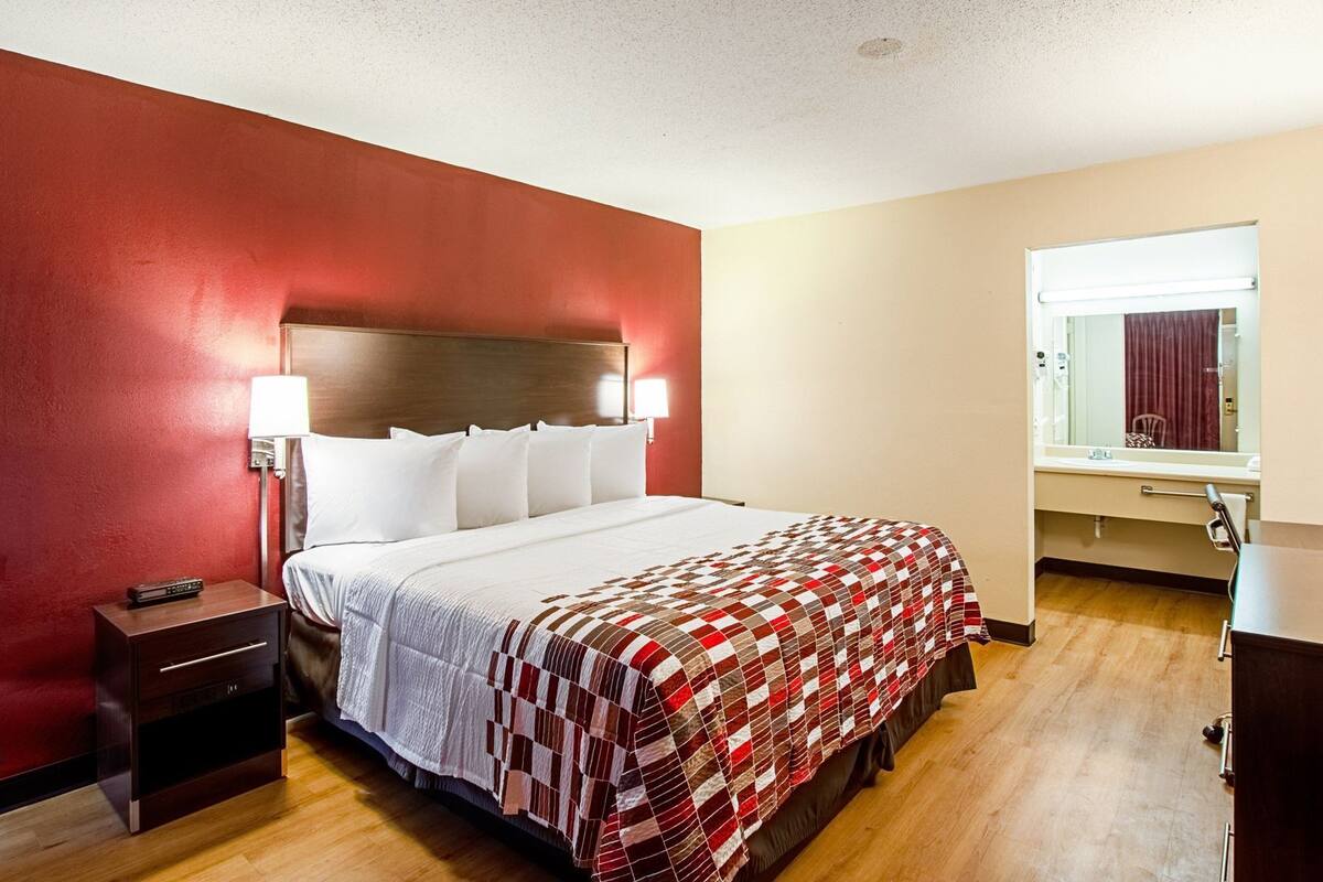 A hotel room with a bed, bedside table, lamp, and a window with a view of a building.