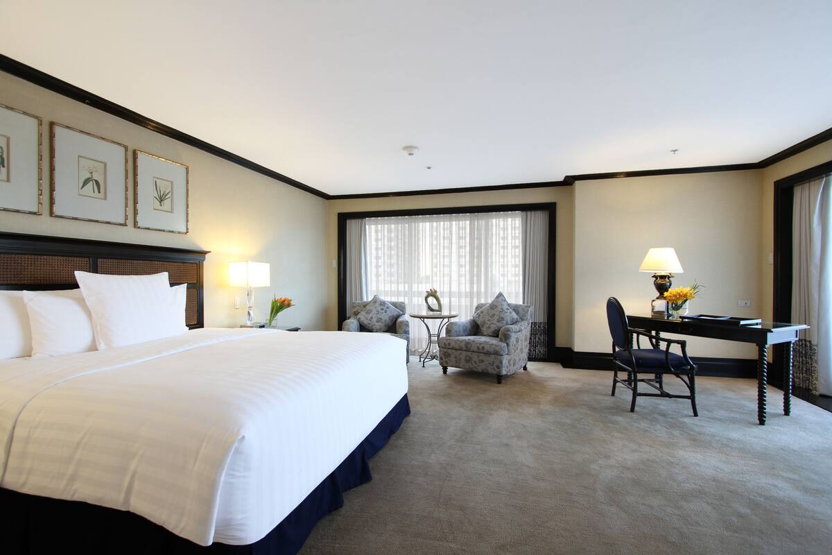 Une chambre d’hôtel avec un grand lit, deux fauteuils, un bureau et une fenêtre avec des rideaux.