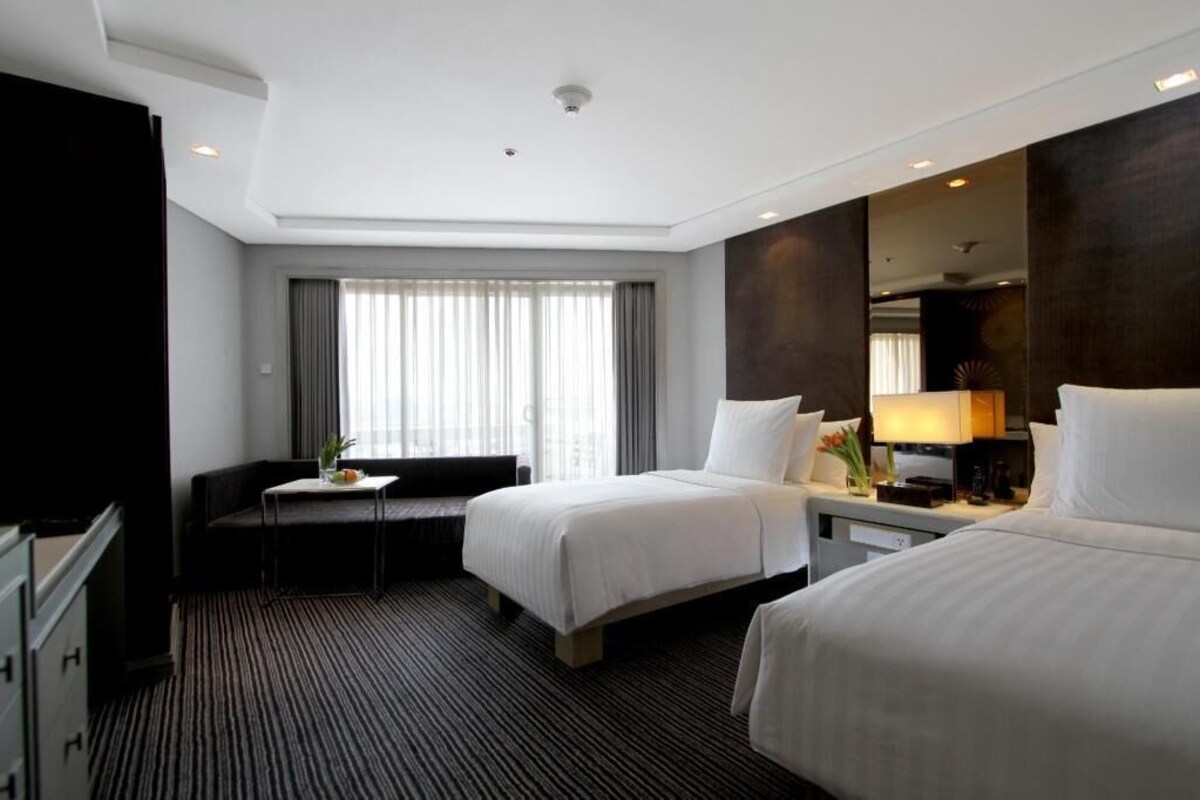A hotel room with two beds, a desk, a chair, a TV, and a large window with curtains.