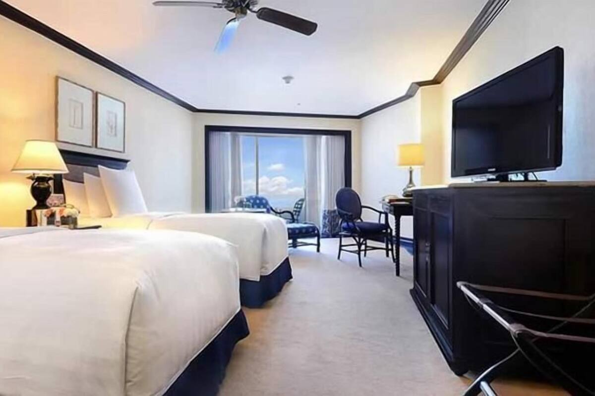 Une chambre d’hôtel avec deux lits, un bureau avec une chaise, une télévision et un ventilateur de plafond.