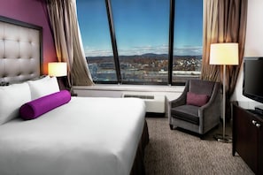 A hotel room with a bed, a chair, a TV, and a large window with a city view.
