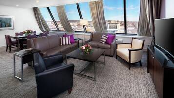 A modern hotel room with a large window, a sofa set, a coffee table, a TV, and a dining area.