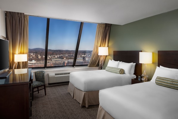 A hotel room with two beds, a desk, a chair, and a large window offering a city view.