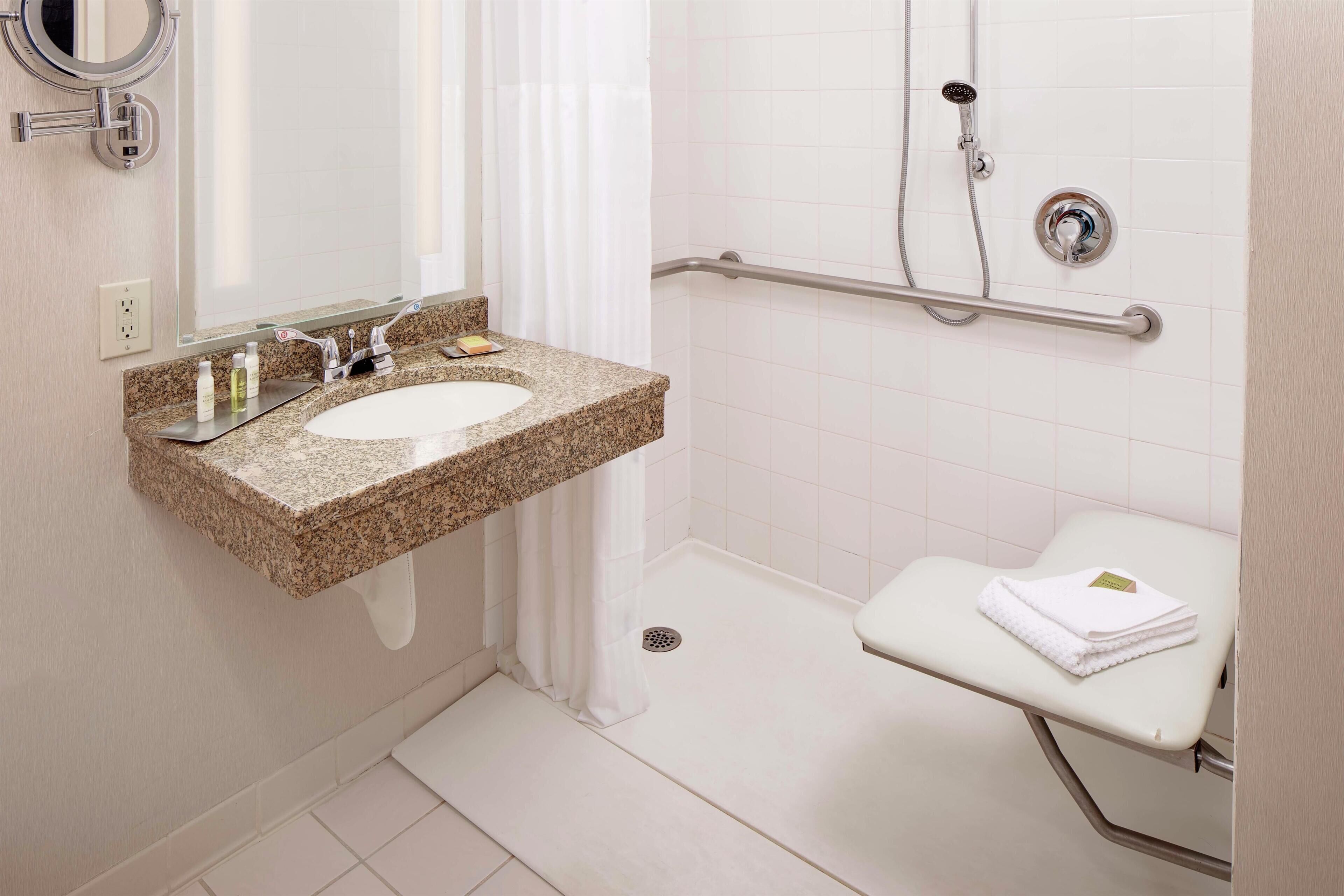 A bathroom with a granite countertop sink, a mirror, a shower with a handheld showerhead, a grab bar, and a white toilet.