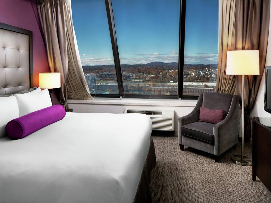 A hotel room with a bed, a chair, a TV, and a large window with a city view.