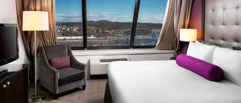 Ein Hotelzimmer mit großem Fenster, einem Bett, einem Sessel, einem Fernseher und einer Lampe.